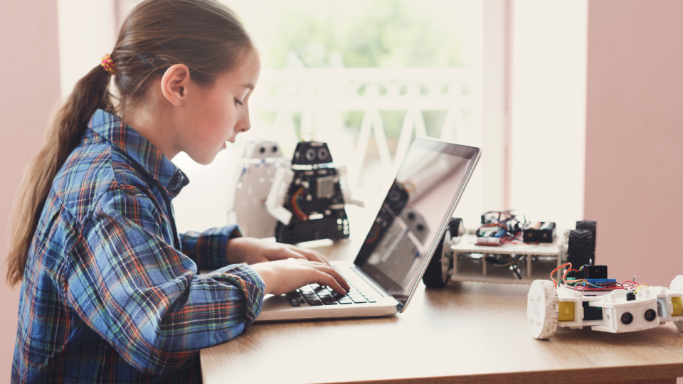 Girl working on a laptop at a desk with several robots around her