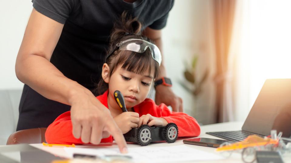 Very young girl using a screwdriver to assemble a robot with the help of an adult
