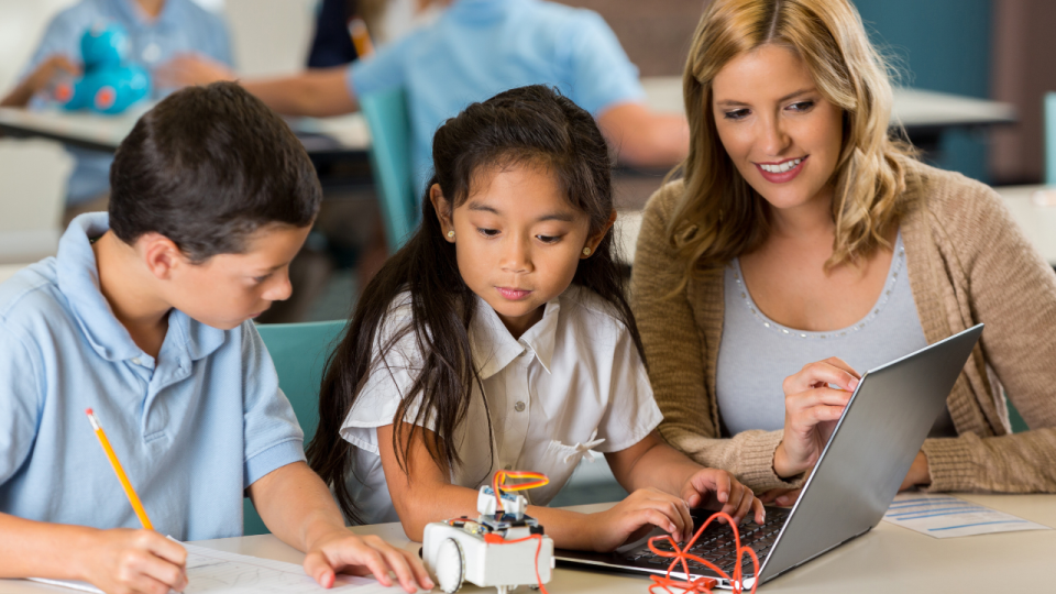 Girl and boy supervised by woman as they program a robot